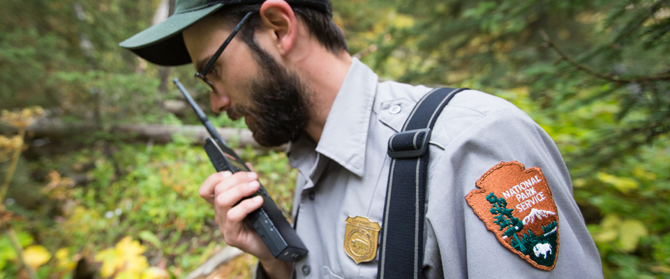 A male park ranger in uniform with an arrowhead patch speaks into a radio A male park ranger in uniform with an arrowhead patch speaks into a radio