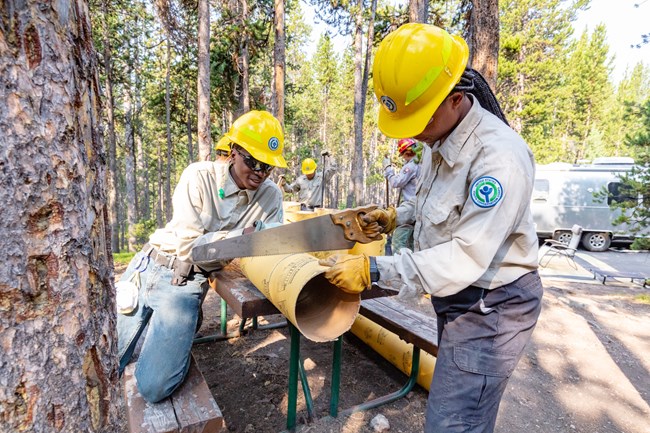two people holding a cutting a tube with a hand saw
