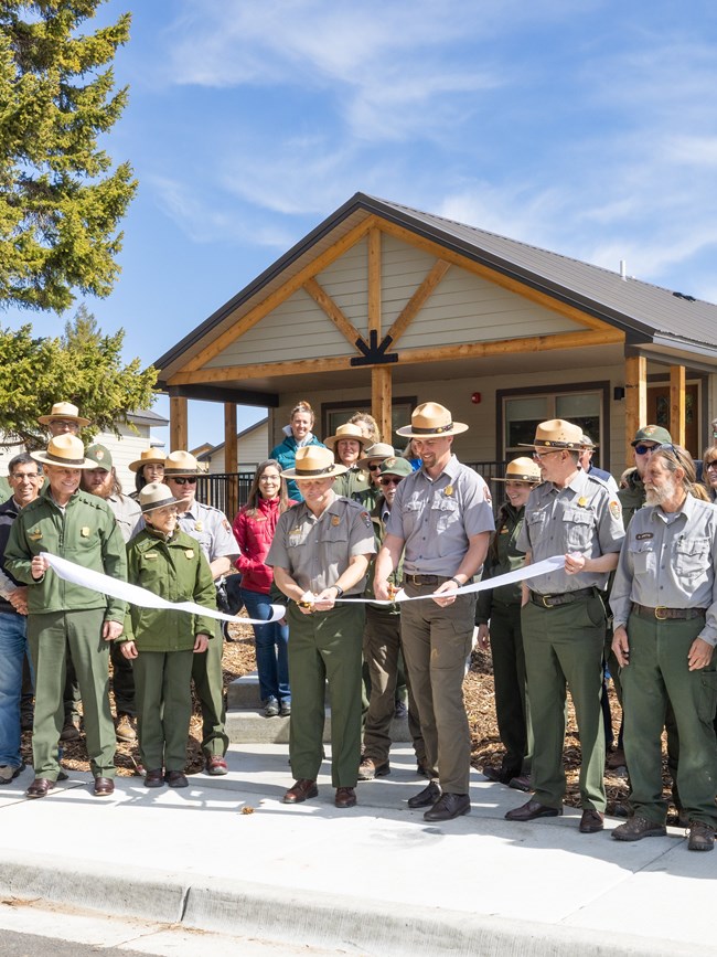 a large group of people cutting a white ribbon in front of new housing