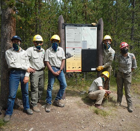 A group of young adults with hard hats stand next to a large sign on a trail.