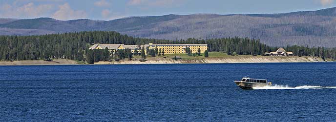A scenic boat cruiser crosses Yellowstone Lake in the foreground. Lake Hotel and mountains are seen in the background. A scenic boat cruiser crosses Yellowstone Lake in the foreground. Lake Hotel and mountains are seen in the background.