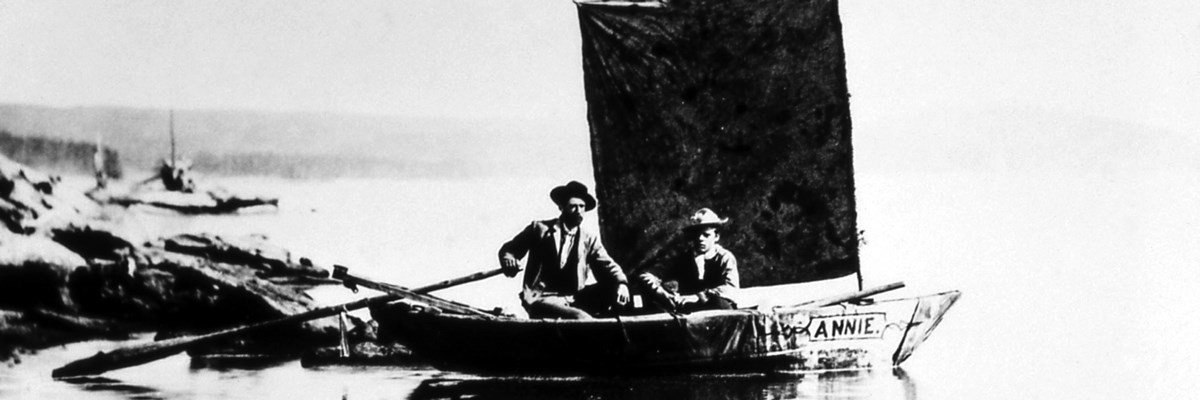 Two men sit on a wooden boat with a square sail Two men sit on a wooden boat with a square sail