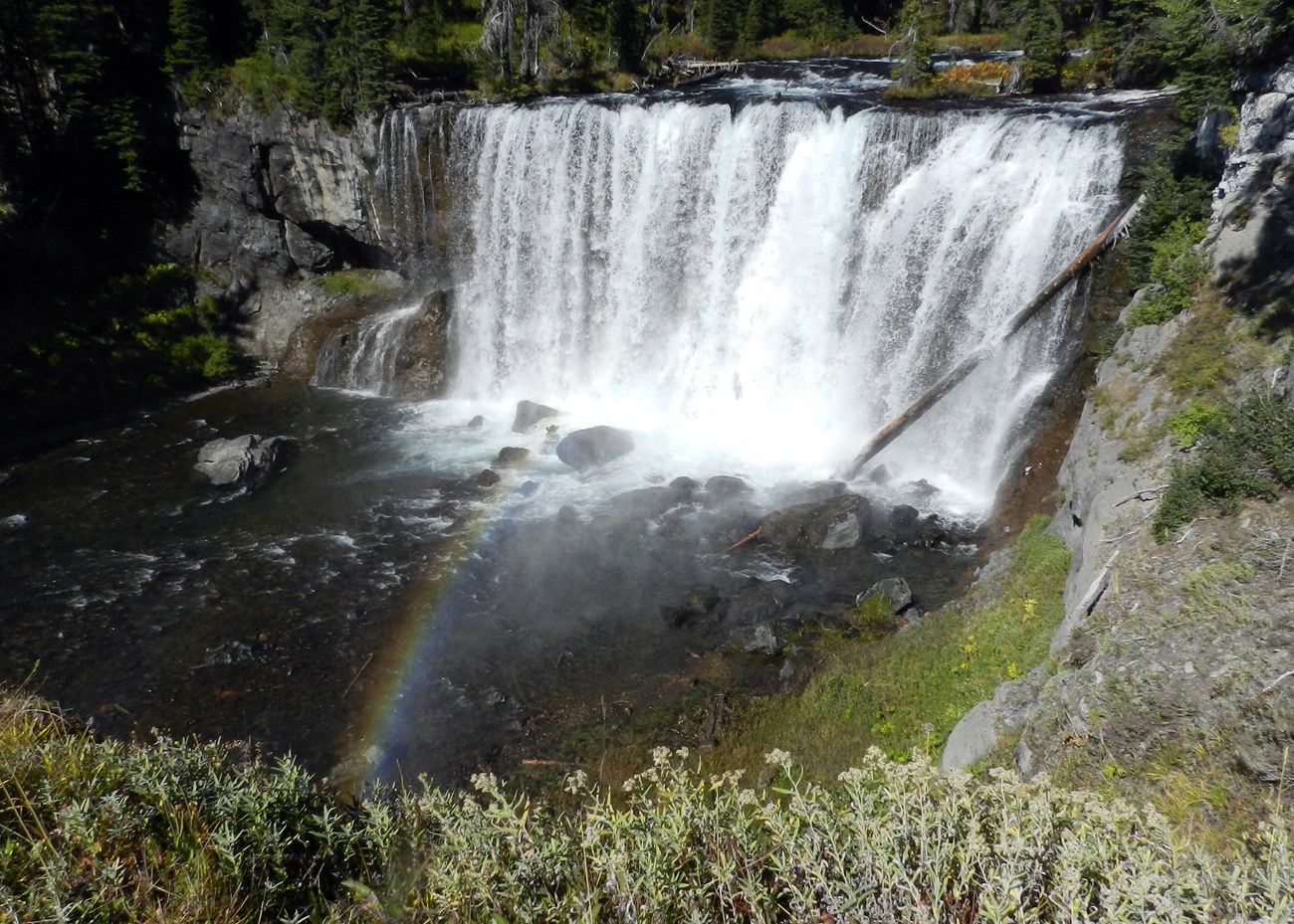 A wide waterfall drops over a gray rock cliff with a rainbow glowing in the spray.