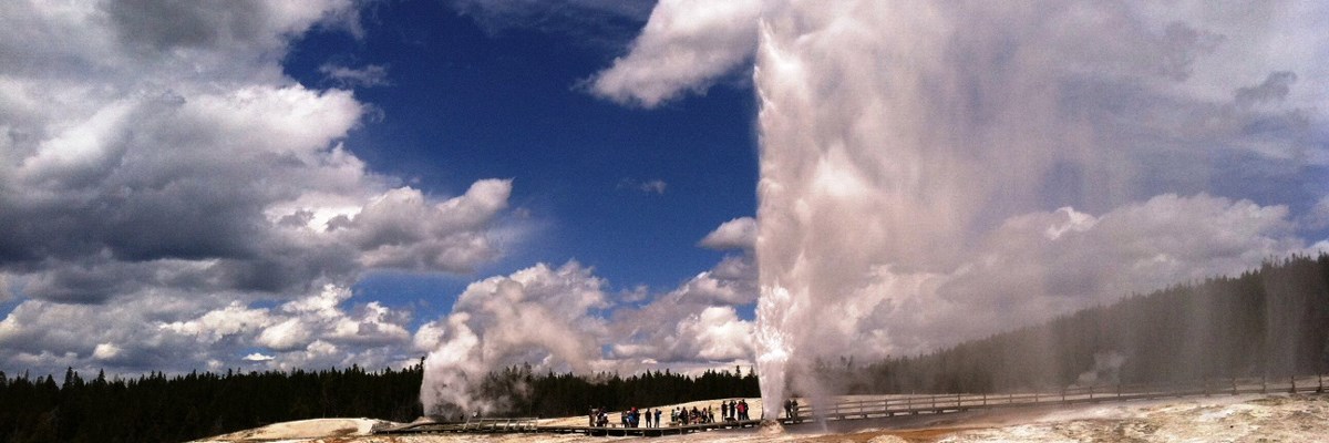Two geysers erupt across a flat, rock landscape with people on a boardwalk Two geysers erupt across a flat, rock landscape with people on a boardwalk