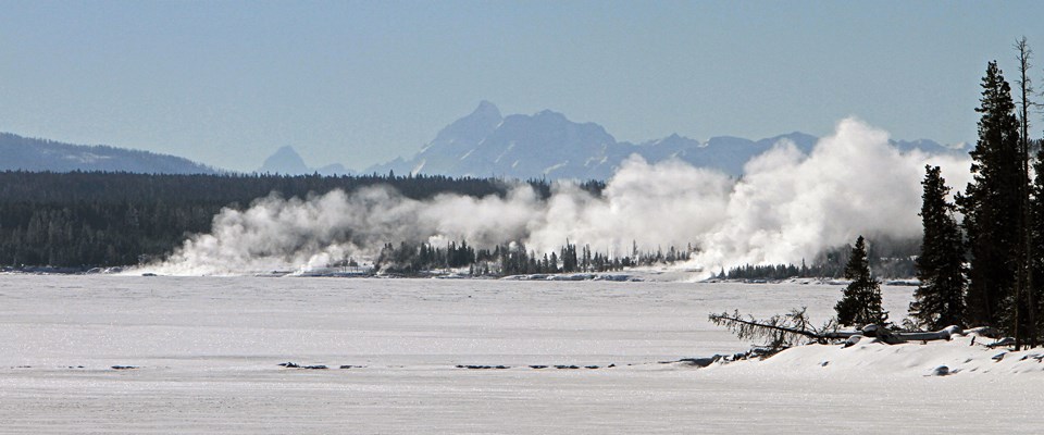 A snow-covered lake with steam rising at the edge and mountains in the background A snow-covered lake with steam rising at the edge and mountains in the background