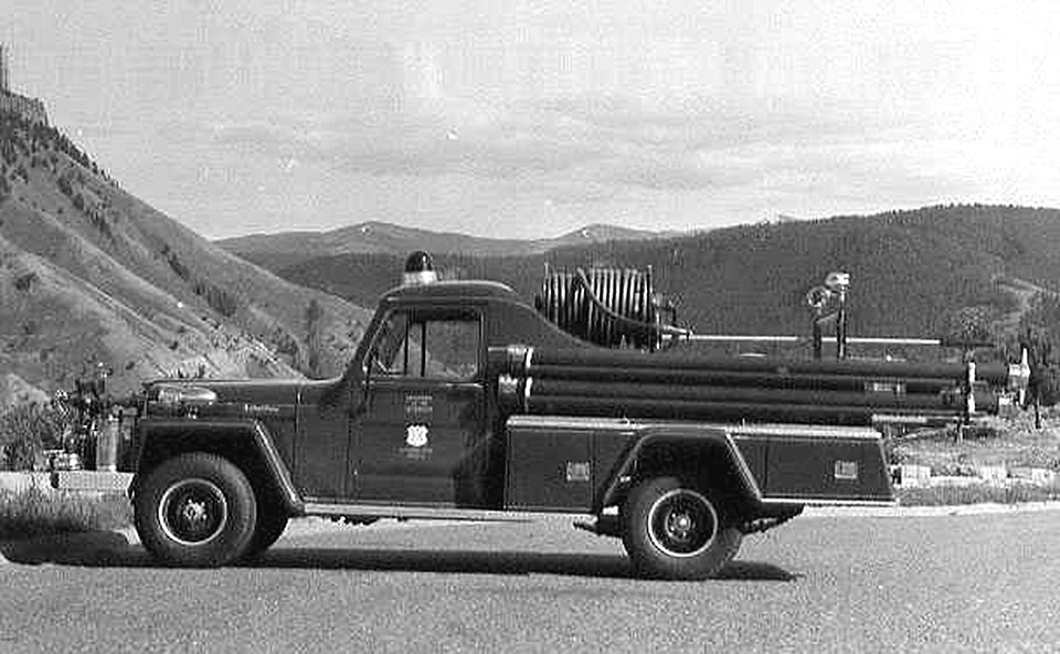 A older style of truck has a fire hose wrapped on a spindle and is parked on the pavement in front of a mountainous and forested landscape.