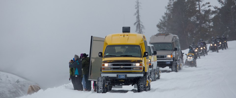 Visitors stand outside of open doors to a van on mattracks with a line of oversnow vehicles and snowmobiles along the side of a road