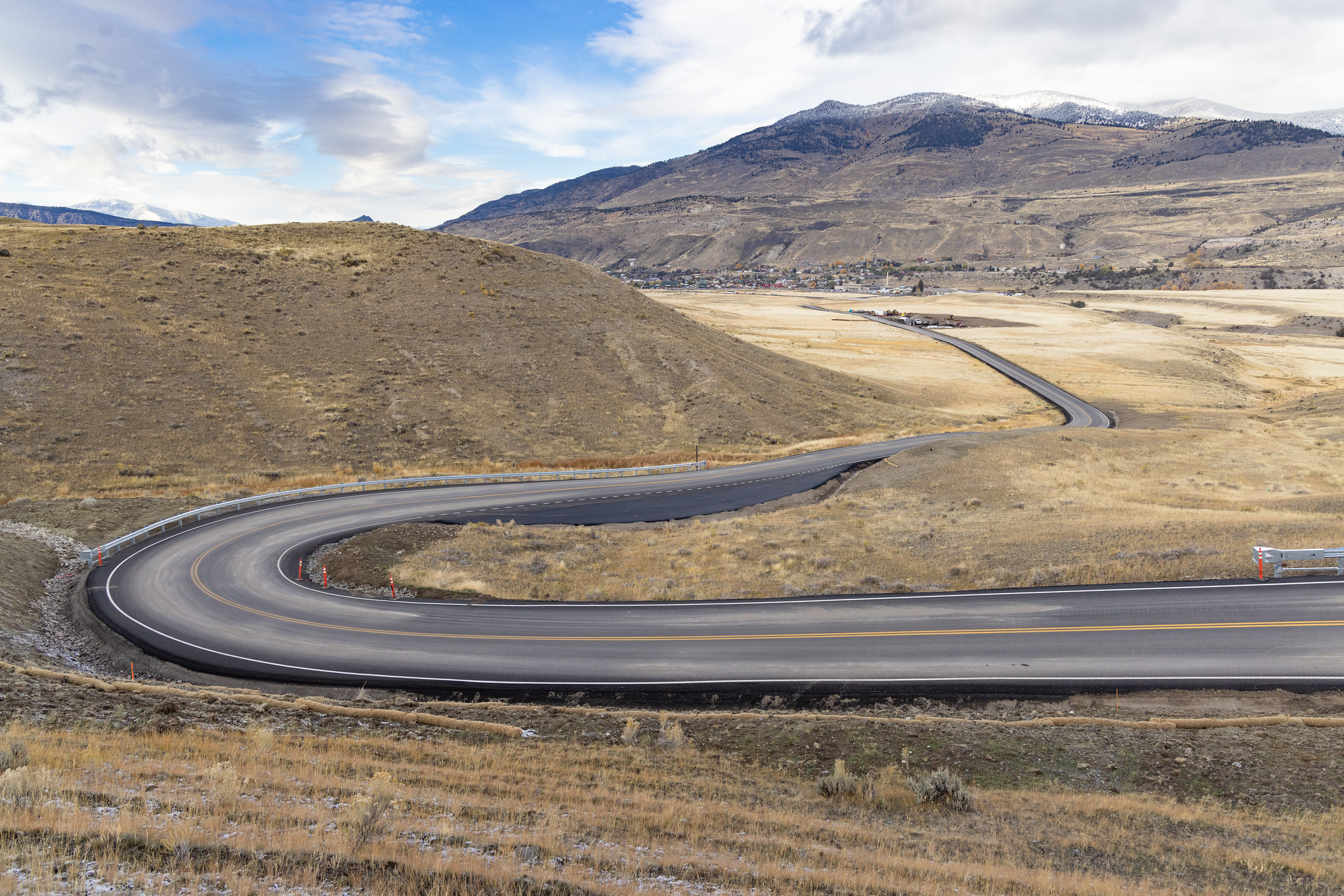 A newly-paved two lane road winds through brown hills with a mountain and a small town in the background.