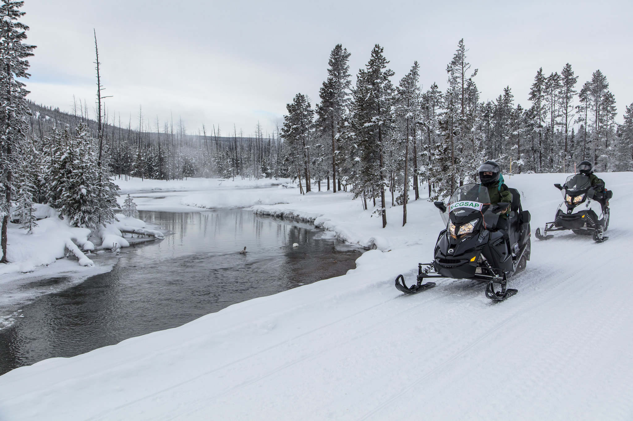 Two people driving snowmobiles on a snowy road near a river