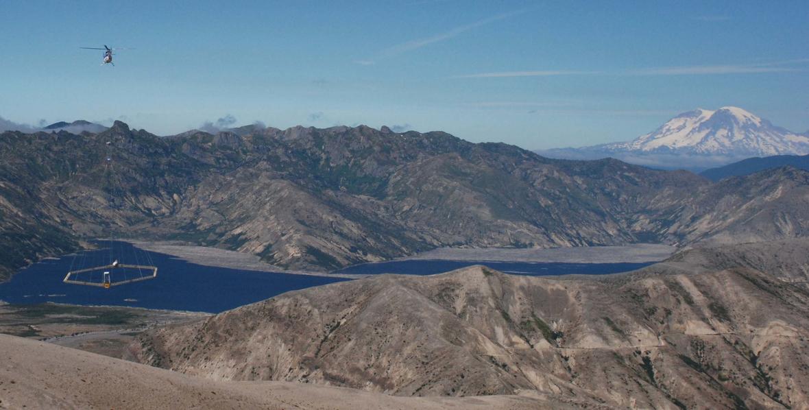 Helicopter flying in a mountain landscape lowering low hanging net over lake.