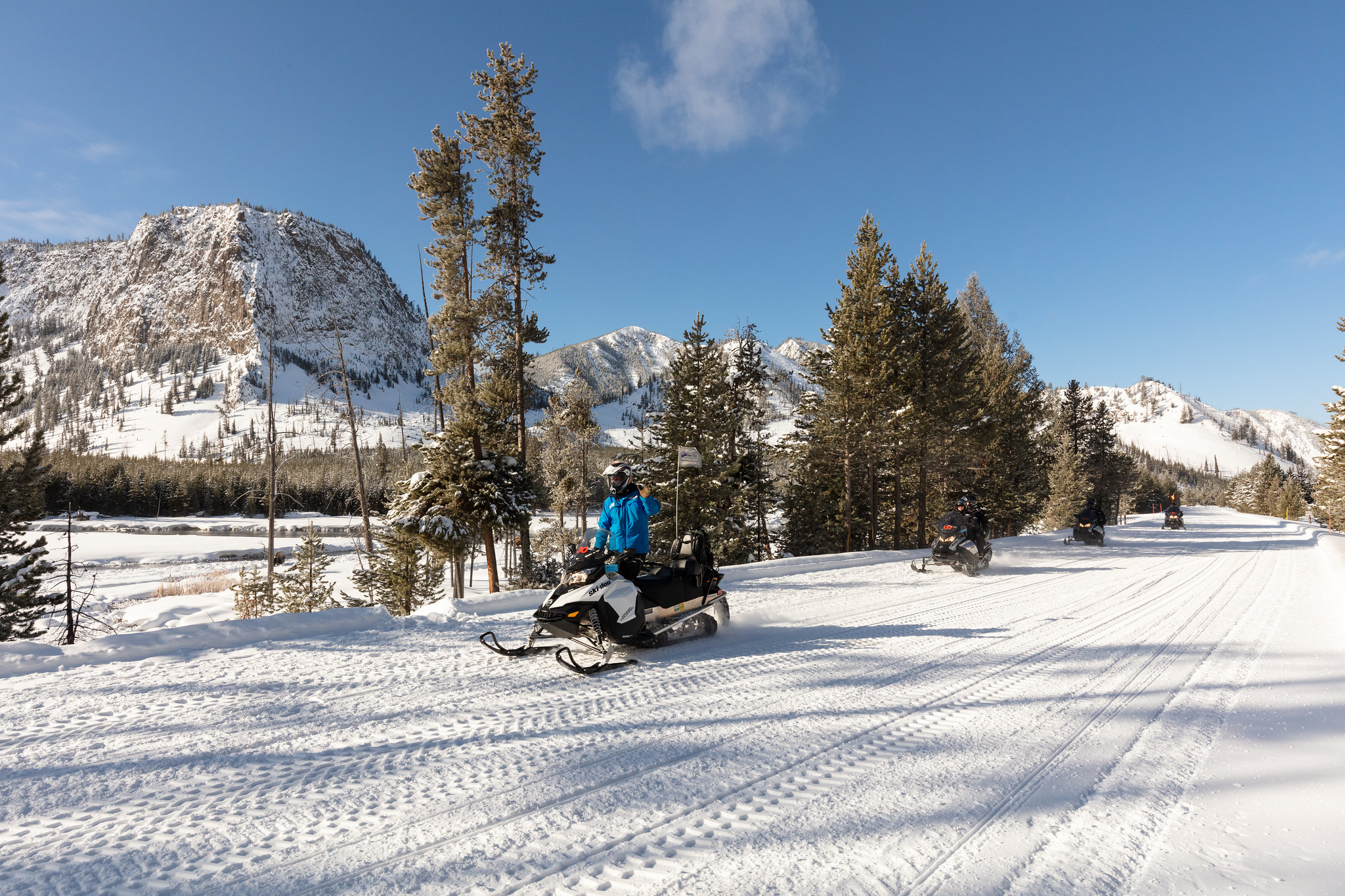 Snowmobile group along Madison River A group of snowmobiles rides on an oversnow road alongside a river.