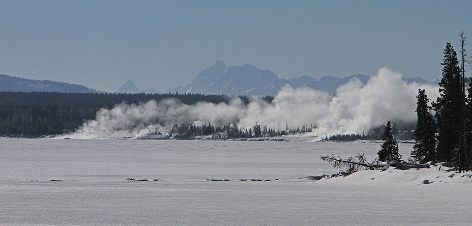 Snowy landscape view across frozen lake with mountain peaks in the distance.