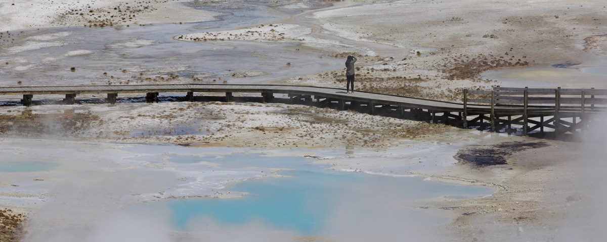 A lone visitor stands on a boardwalk and photographs the nearby hot springs.