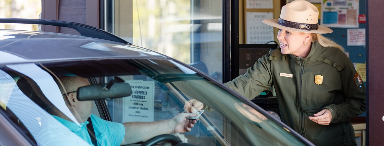 A female park ranger hands a visitor in a car a park guide at an entrance station.