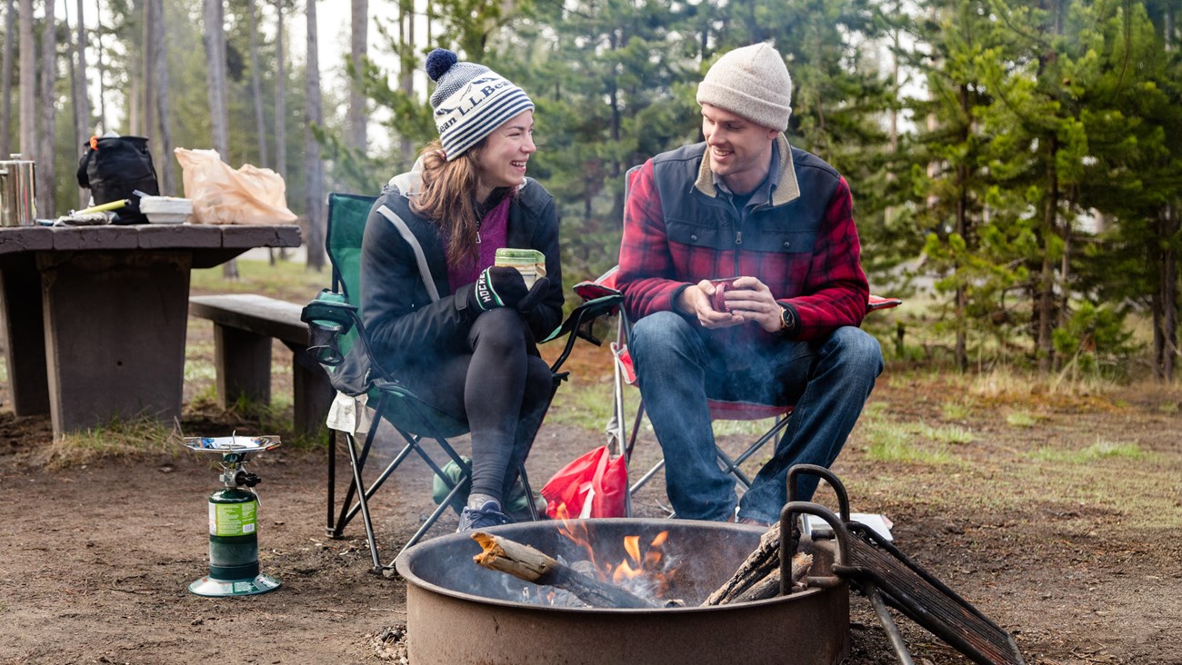 Campers sit by a fire with hot drinks