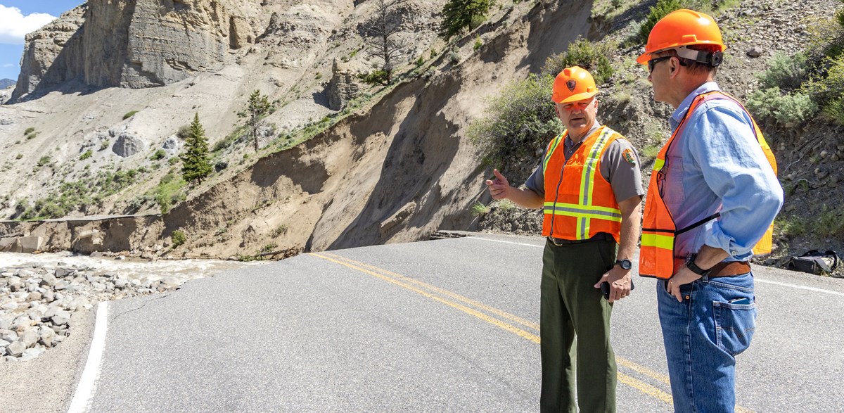Two men wearing hard hats talk while surveying a section of mountainous road that has eroded into the river below.