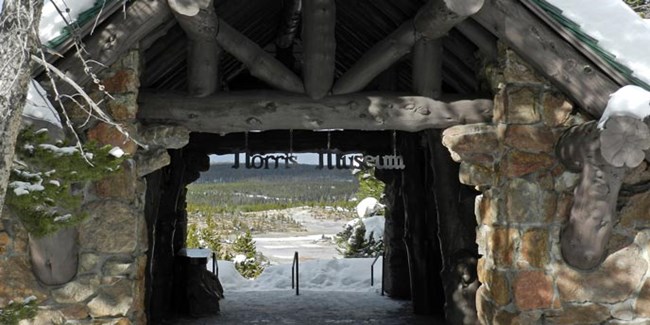 A metal sign reading "Norris Museum" hangs in a log entry way with a view of a geyser basin