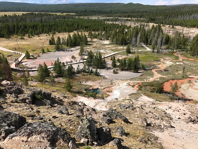 View from the top of a hill showing a boardwalk trail leading past steaming hot springs.