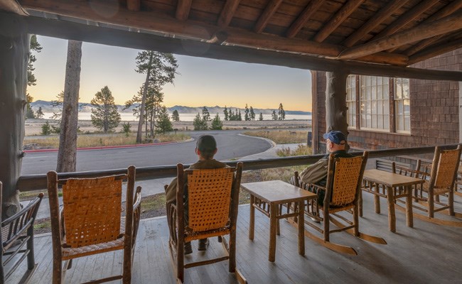 Two men sitting in rocking chairs of a rustic porch, looking out over a large lake.