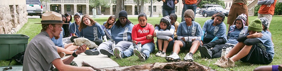 Male ranger sits on a sidewalk with animal pelts and talks with a large group of teenager, many of which are also seated on the ground.