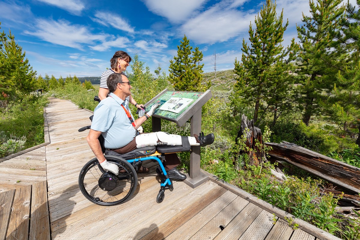 Two people, one in a wheelchair, read a wayside exhibit on a boardwalk