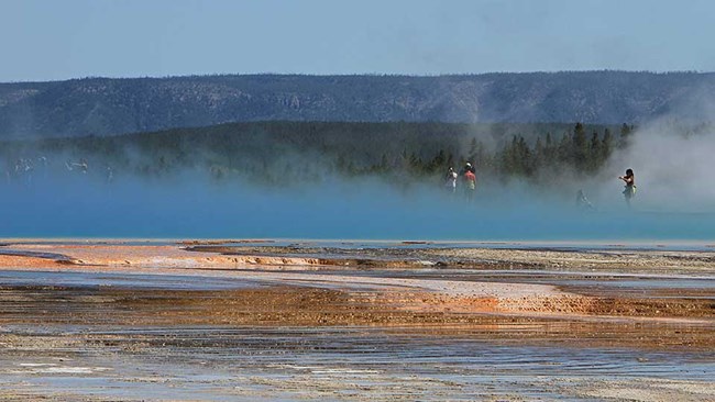 Blue steam rises from a blue hot spring, with a few visitors on a boardwalk visible through the steam.