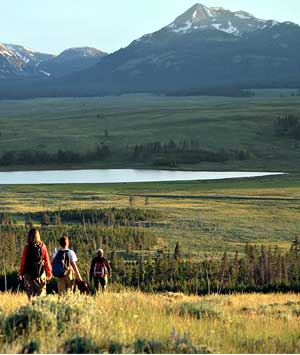 Three hikers walk down the trail toward a lake and mountain peaks in the background. Three hikers walk down the trail toward a lake and mountain peaks in the background.
