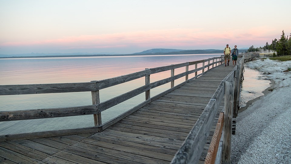 Two people walking on a wooden boardwalk next to a lake at sunset.