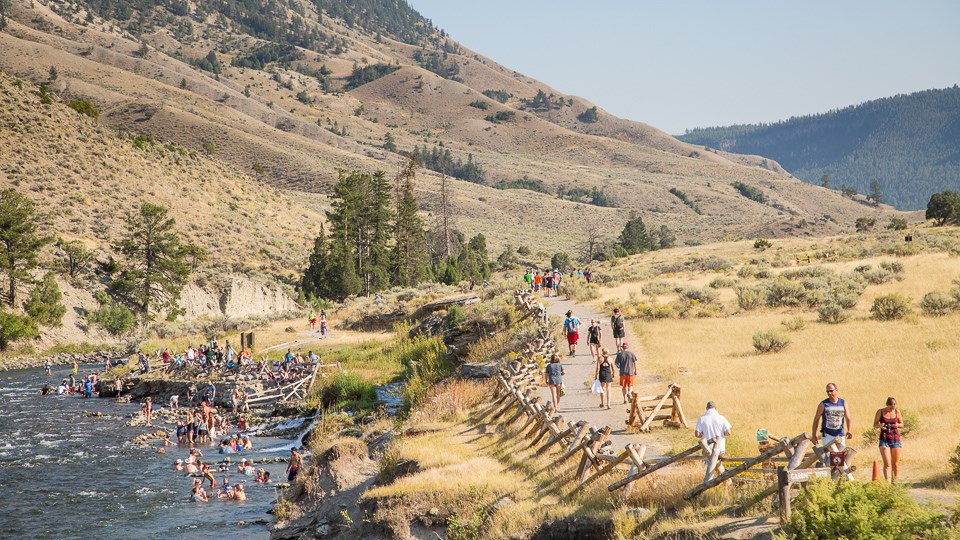 A crowd of people sit in a river while others stand on shore or walk along a trail next to the river. A crowd of people sit in a river while others stand on shore or walk along a trail next to the river.