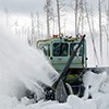 A mint green colored rotary snow plow cuts through piles of snow.