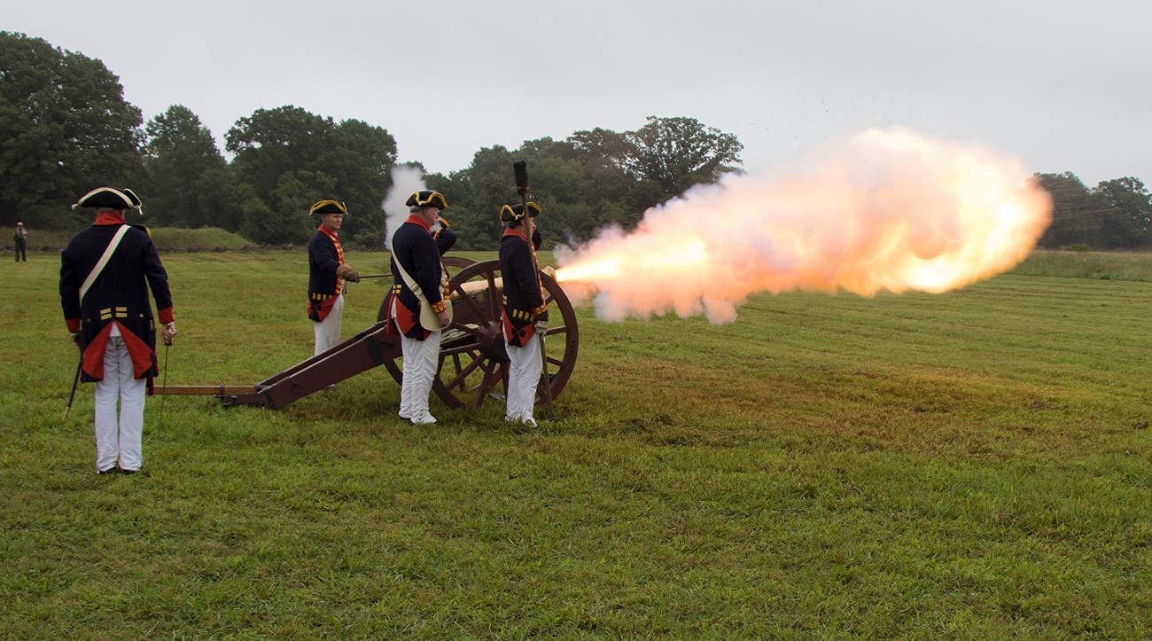 Cannoniers in blue Royal Artillery coats fire a brass cannon with a fire ball shooting out of the muzzle