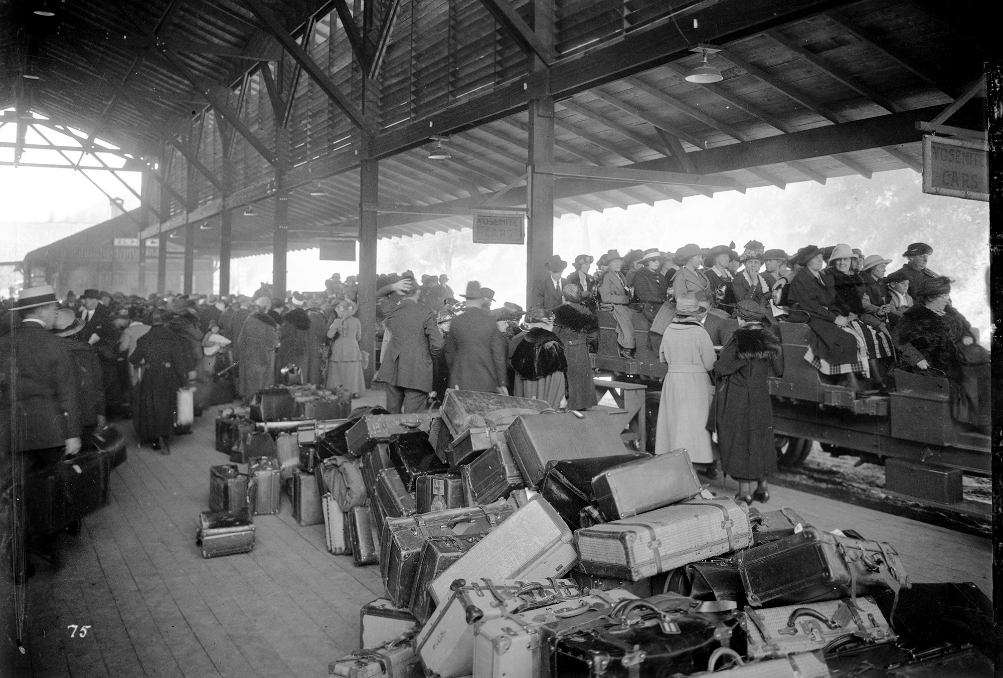 A black and white photo of a crowded train platform covered in suitcases, with passengers loaded onto 1910s-style buses
