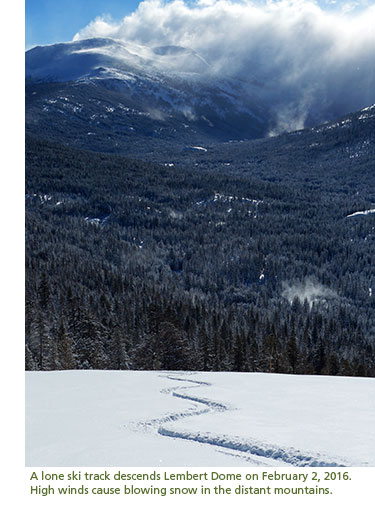 A ski track descends a hill while snow blows in the wind on distant mountain tops.