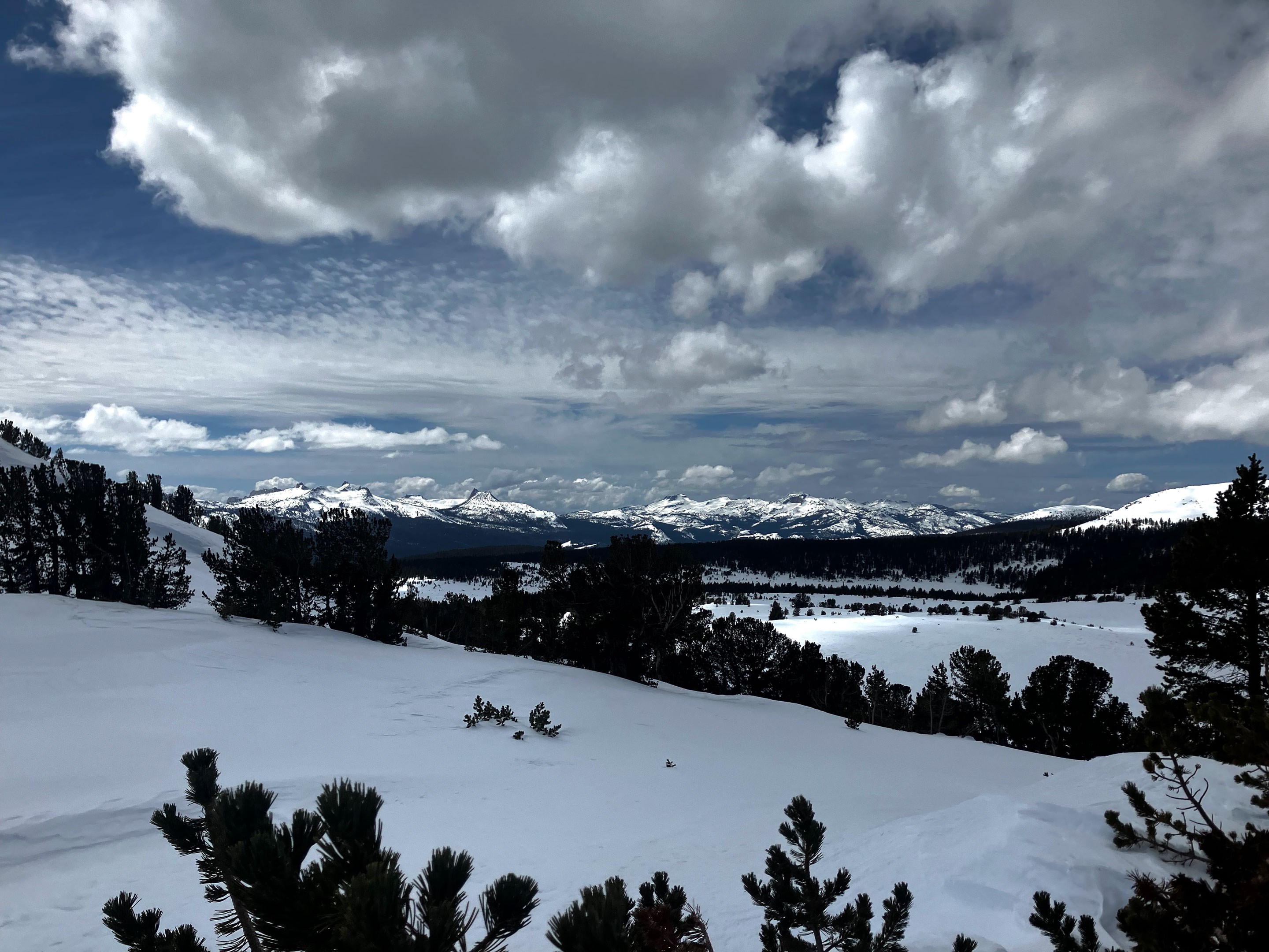 Sanding on a snowy hill will rolling snowy hills in foreground and snowy peaks in background; partly cloudy sky with cumulus, altostratus, and altocumulus clouds