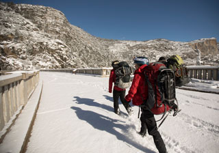 Hikers walking across snow-covered dam toward hills Hikers walking across snow-covered dam toward hills
