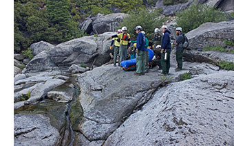 Several rescuers surround patient in a sleeping bag lying down in a litter  near a creek