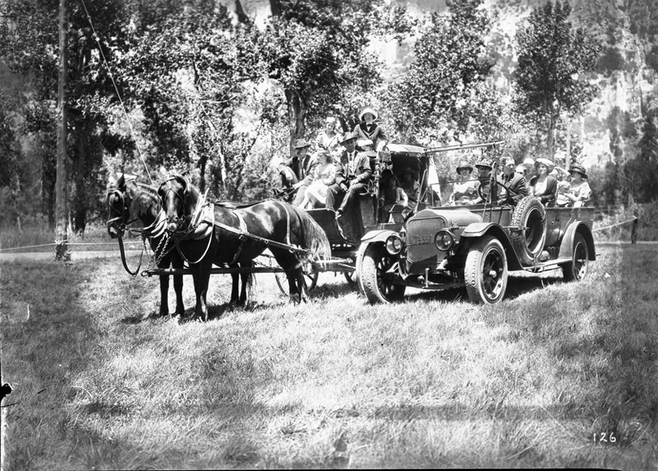 People sitting on a horse-drawn stage next to a touring car filled with people