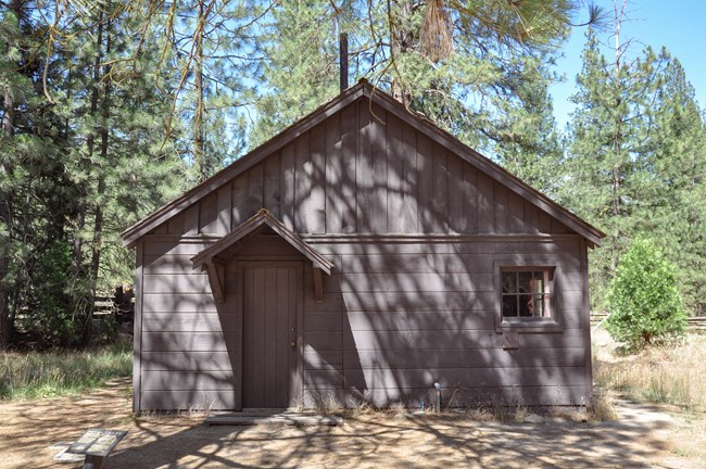 Los árboles rodean una sencilla cabaña de madera que tiene una puerta y una pequeña ventana cuadrada en el frente.