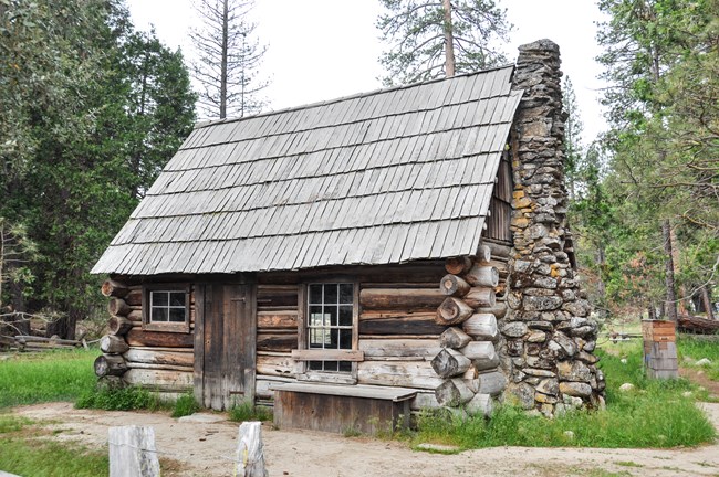 La hierba crece alrededor de una pequeña casa de troncos que tiene una chimenea de piedra.