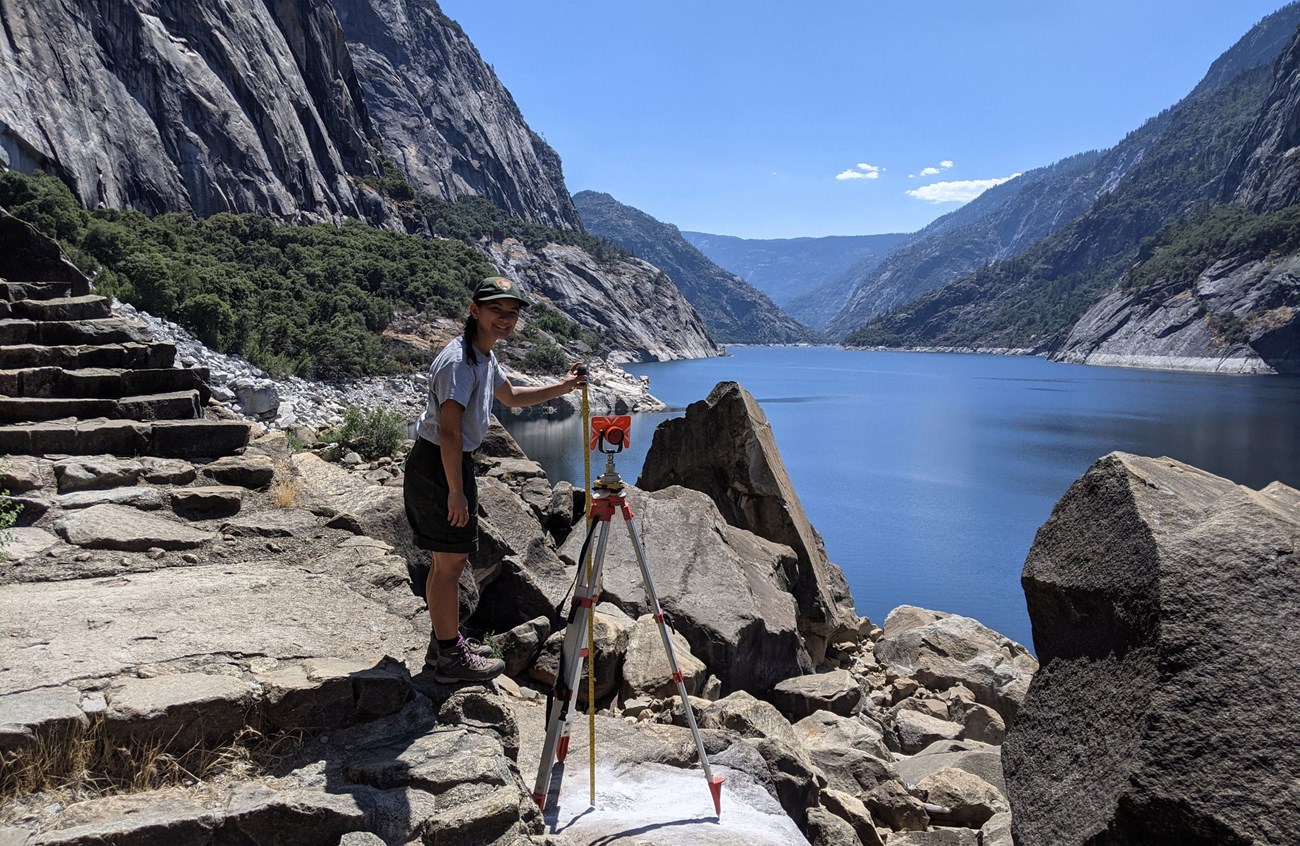 Engineering intern setting up survey equipment at Wapama Falls at Hetch Hetchy with reservoir and granite peaks in the background.