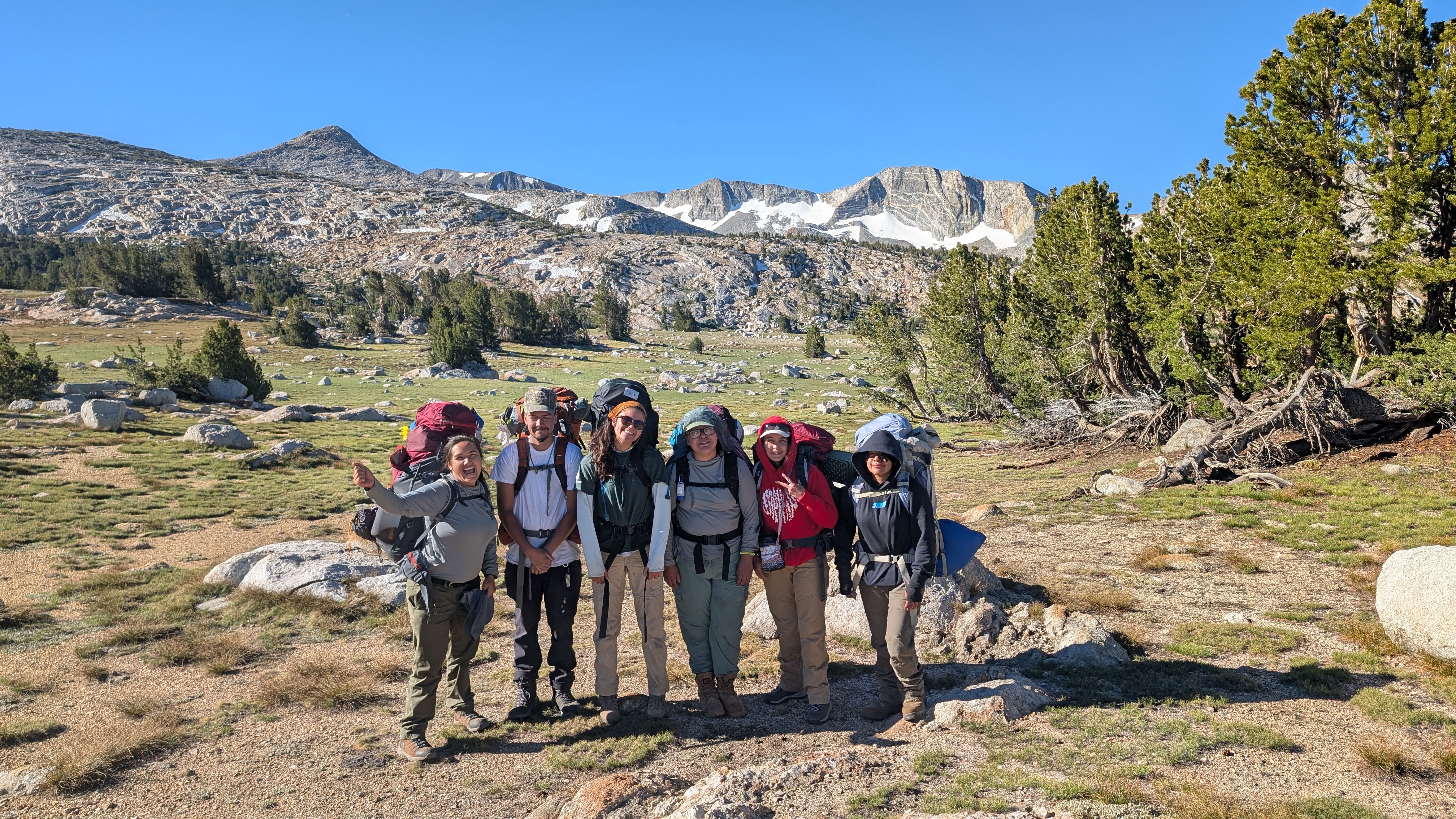 A group of six people with backpacking gear pose in front of a mountain range in Yosemite National Park.