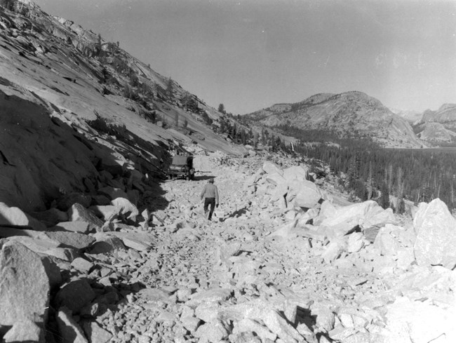 A worker walks through rubble along a newly blasted section of granite; unblasted granite is seen upslope and a tall granite dome in the distance