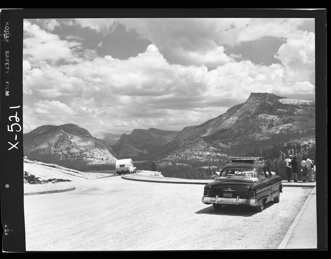 A paved pull-out area alongside a road with a car parked, a car driving pulling a trailer, and tourists gathered at an interpretive sign; peaks and domes are in the distance