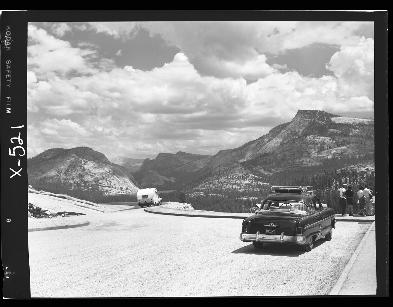 Olmsted Point on Tioga Road A paved pull-out area alongside a road with a car parked, a car driving pulling a trailer, and tourists gathered at an interpretive sign; peaks and domes are in the distance