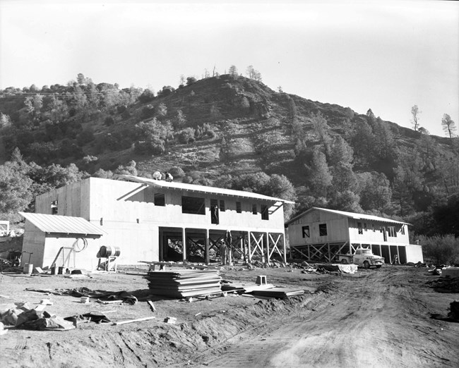 Two homes partially constructed on a hillside; men are seen working on the roof and on the framing below; construction materials and a work truck are scattered in the area