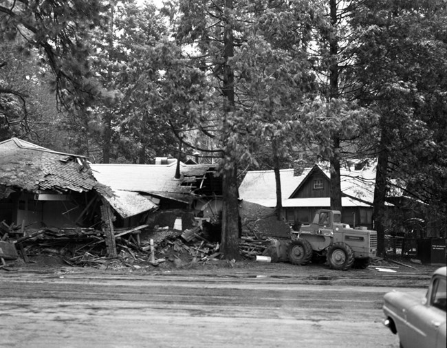 Several buildings being razed: they are partially collapsed with a bulldozer working in the area and several mature pine trees