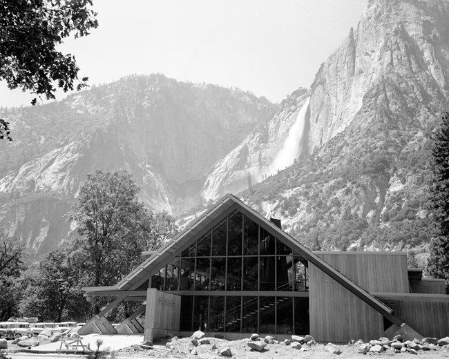 A-frame building with a large wall of windows and wood paneling; a concrete sign with lettering "Degnan's" is visible in front; cliffs and a large waterfall are behind it