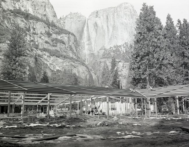Steel framing begins to take the shape of a building; a crane is working in the area and two men sit on the roof framing; there is snow on the ground and Upper Yosemite Falls is slightly flowing in the background