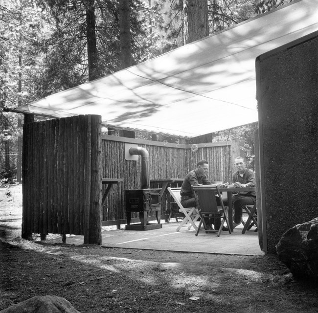 Two men sit at a round table underneath a canvas shade; the paved area is partially fenced in with wood branches and logs