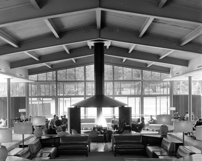 A lounge with a fireplace at center and ample lounge furniture; the high ceiling has exposed beams and the opposite wall is all windows